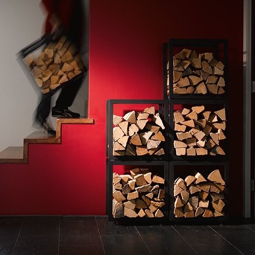 Mann trägt Feuerholz neben Holzregal vor roter Wand